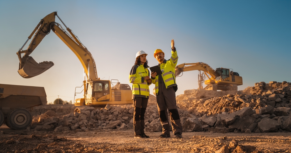 Hispanic Female Inspector Talking to Caucasian Male Land Development Manager With Tablet On Construction Site Of Real Estate Project. Excavators Preparing For Laying Building Foundation. Hot Sunny Day