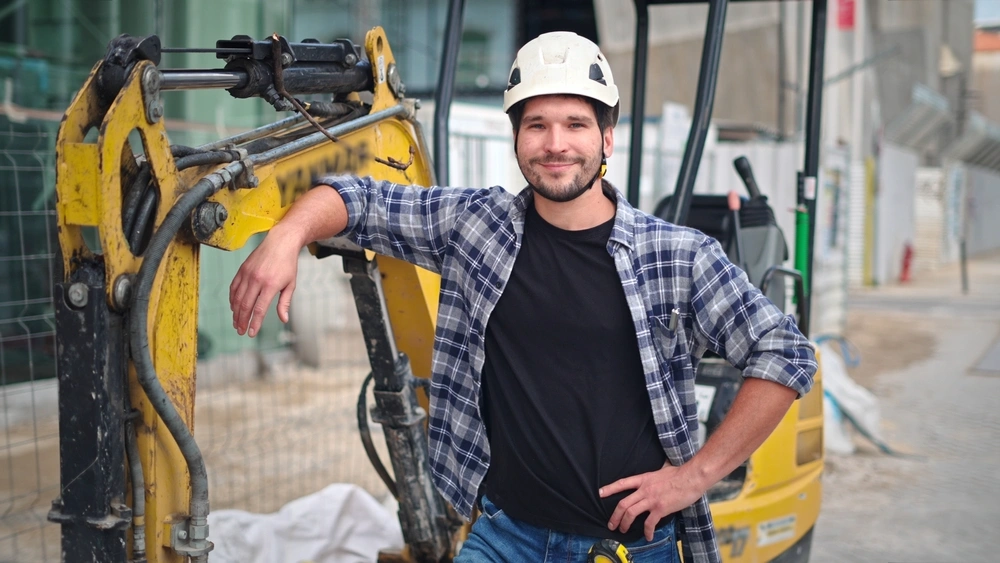 Confident construction site worker portrait. Smile constructor man look camera. Builder engineer wear white helmet. Excavator driver plaid shirt. Guy build new house city street. Erector person job.