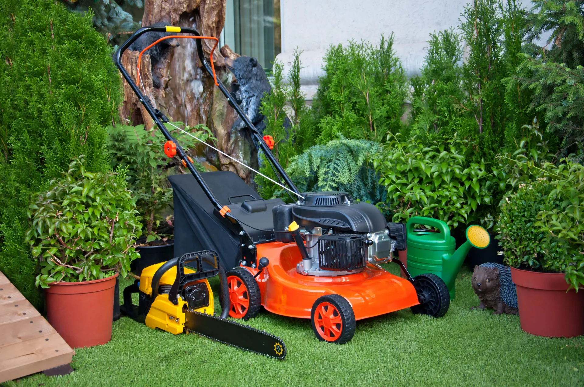 Gardening equipment, orange lawnmower, yellow chainsaw, green watering can, garden statue of a hedgehog, garden plants in pots in the garden